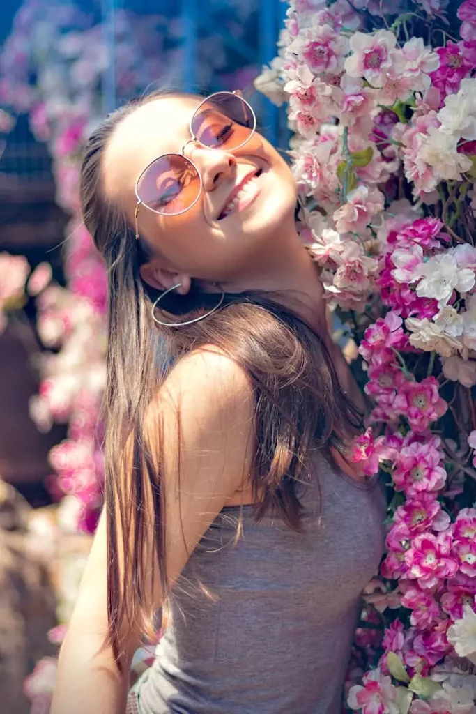 Joyful young woman with sunglasses posing with vibrant pink flowers outdoors.